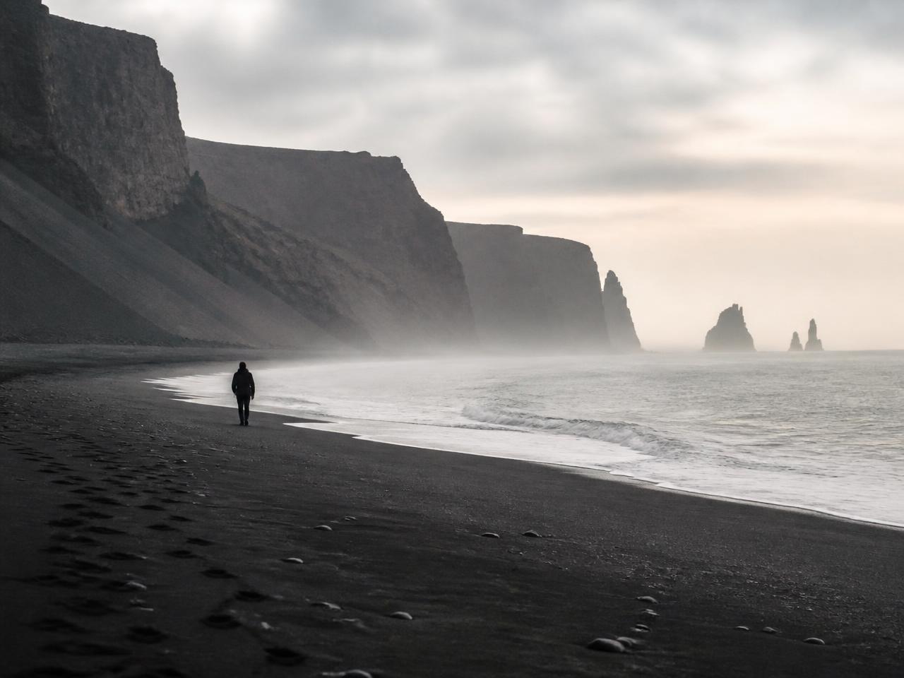 A lone figure walking a misty dark sand beach beneath tall cliffs